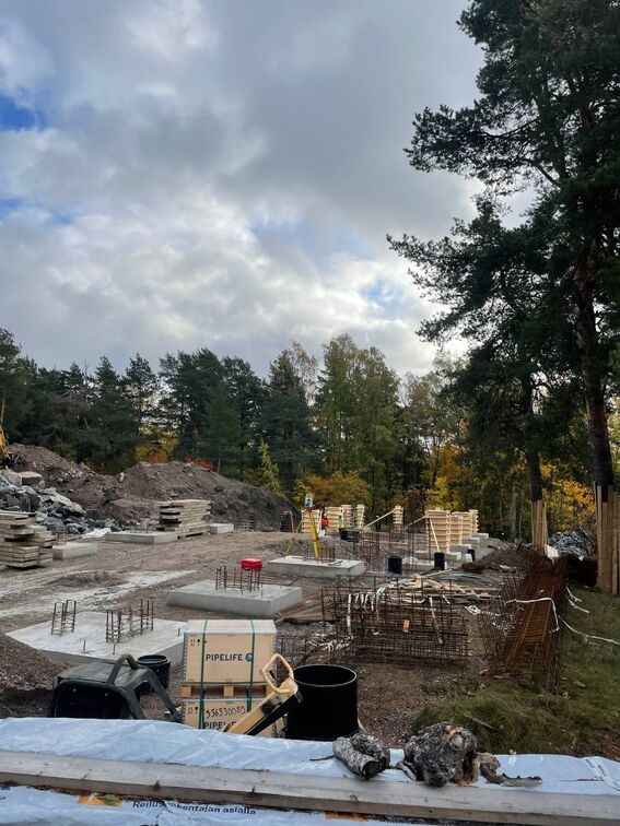 Construction site with various building materials and equipment, surrounded by autumn trees under a cloudy sky.
