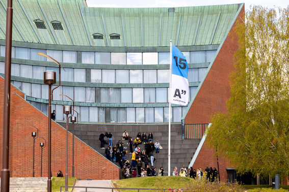 Aalto 15 years blue flag floating in front of outside amphitheatre. A group of people are gathered on the steps.