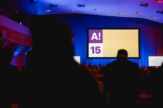 Audience in a hall watching a large screen displaying 'A! 15' with blue curtains and a wooden ceiling.