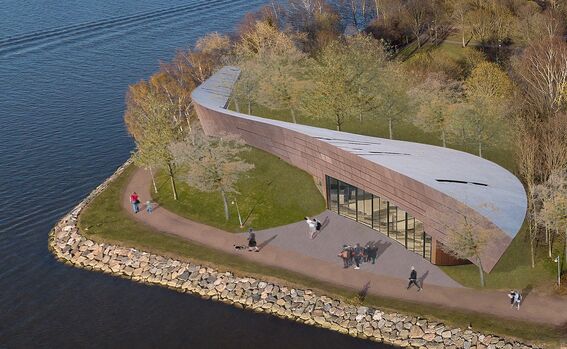 A modern, curved building with a silver roof by a lakeside. People walk on a pathway surrounded by trees.