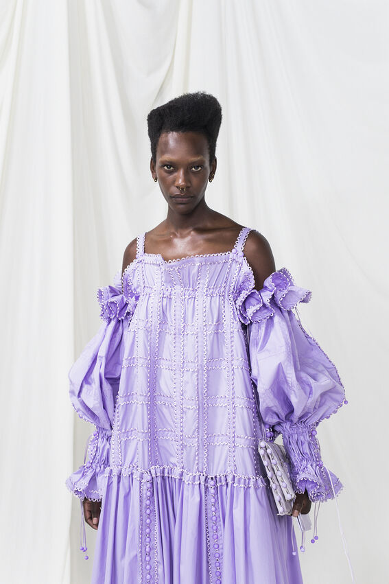 Person wearing a light purple dress with puffed sleeves and intricate patterns, holding a matching clutch.
