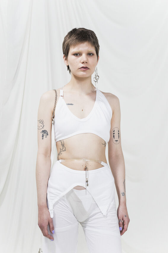 Person with short hair wearing a white crop top and pants, with multiple tattoos on arms and torso.