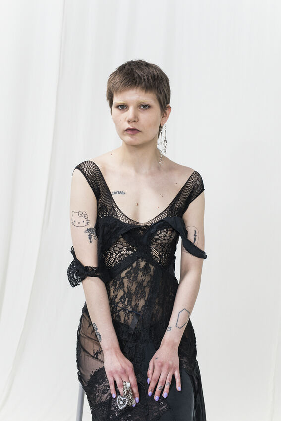 Person with short hair wearing a black lace dress, showing tattoos on arms and chest, sitting against a white background.