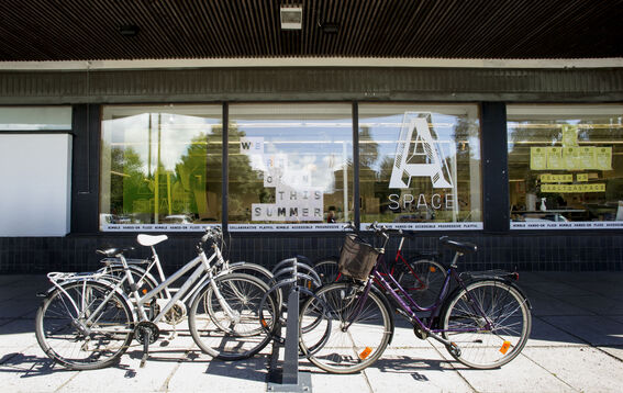Several bicycles are parked in front of a building with large windows displaying signs about summer opening.
