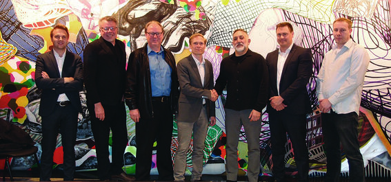 Six men in formal attire stand in front of a colourful abstract mural. Two men in the centre are shaking hands.