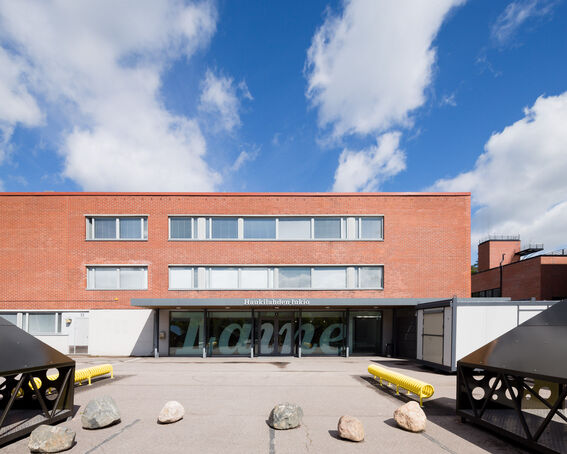 A red-brick building with large windows and a sign reading 'Haukilahden lukio'. The sky is blue with scattered clouds.