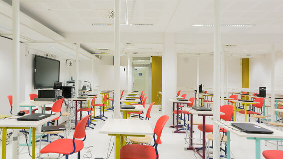 A modern classroom with red chairs, white tables, and computers. A large screen is mounted on the wall.