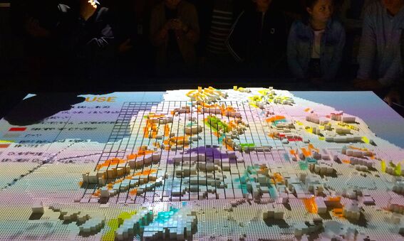 A 3D map with colourful blocks on a table, surrounded by people observing it.