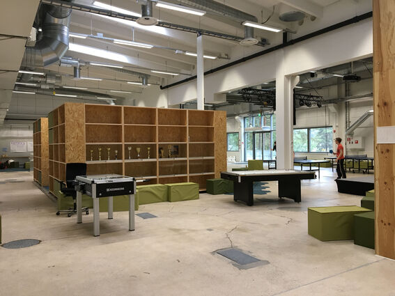 Spacious room with wooden shelves, table football, green seating, and people playing table tennis near large windows.