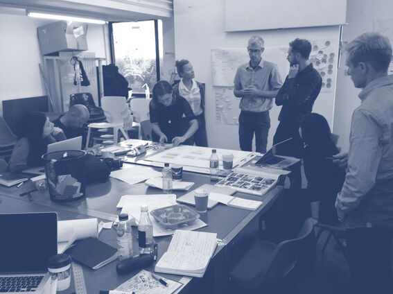 A group of people working around a table with laptops, papers, and drinks in an office setting.