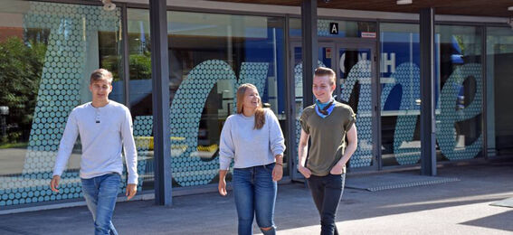 Three people walking outside a building with large windows and hexagonal patterns. The word 'love' is visible on the glass.