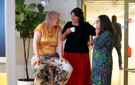 Three adults chatting indoors; one seated, two standing with a coffee cup, in colourful clothes by a tall plant.