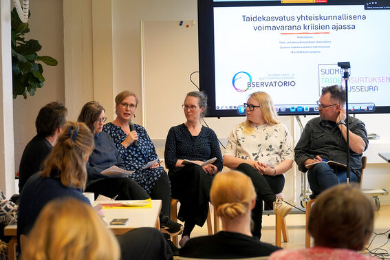 Panel of adults discussing art education in a classroom, audience seated, presentation slide on large screen.