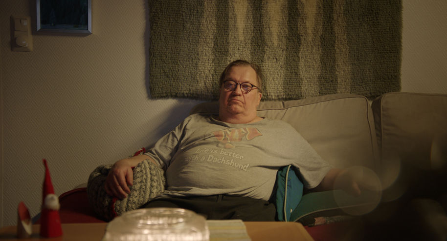 vakavan näköinen vanhempi mies istuu kotisohvalla ja katsoo kameraan