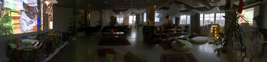 Tunnelmallisesti sisustettu luokkahuone valoineen ja lattiaistuimineen. Hämärä valaistus, videotykin heijastama lämmin kajo mehiläistarhauksesta.