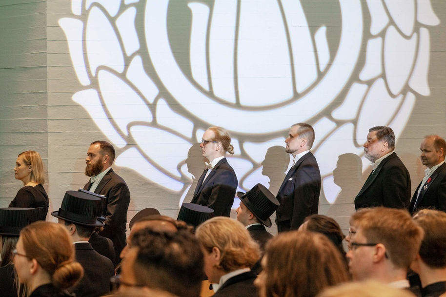 Uudet tohtorit jonottavat lavalle. Taustalla Tiedon liekki heijastettuna valkoiselle seinälle.