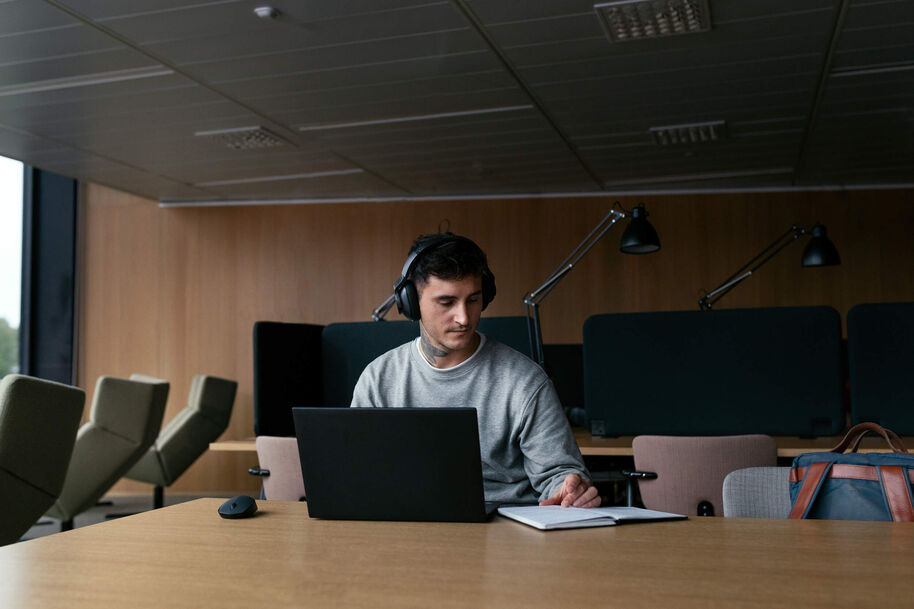 Male student with a large tattoo on his neck looks at his laptop while making notes in a notebook