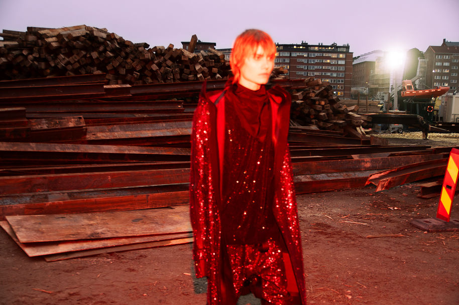 Henkilö punaisessa strassein koristellussa takissa kävelee rakennustyömaalla, jonka taustalla on puupinoja.