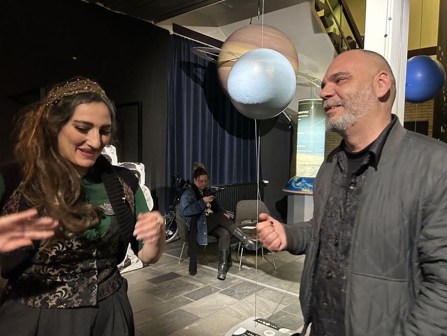 Two people indoors near hanging planet models. A seated person is in the background using a phone.