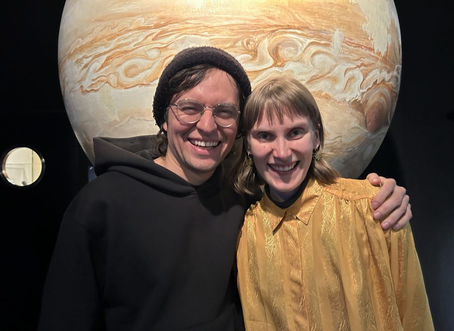 Two people standing in front of a large planetary model. One is wearing a black hooded sweatshirt and the other a yellow blouse.