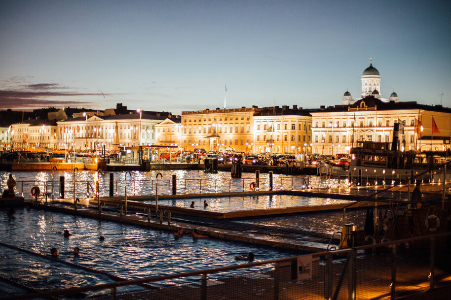 Night view of Helsinki's waterfront with swimming pools in foreground, illuminated buildings and cathedral in background.