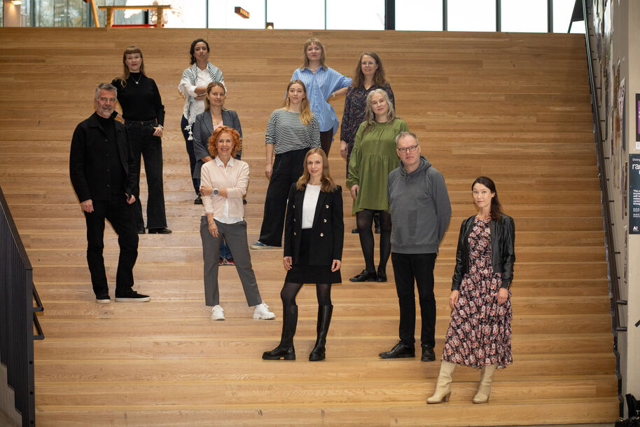 A group of people standing on wooden steps indoors. They are wearing casual and semi-formal clothing in various styles. Group is of Creative Leap research team.