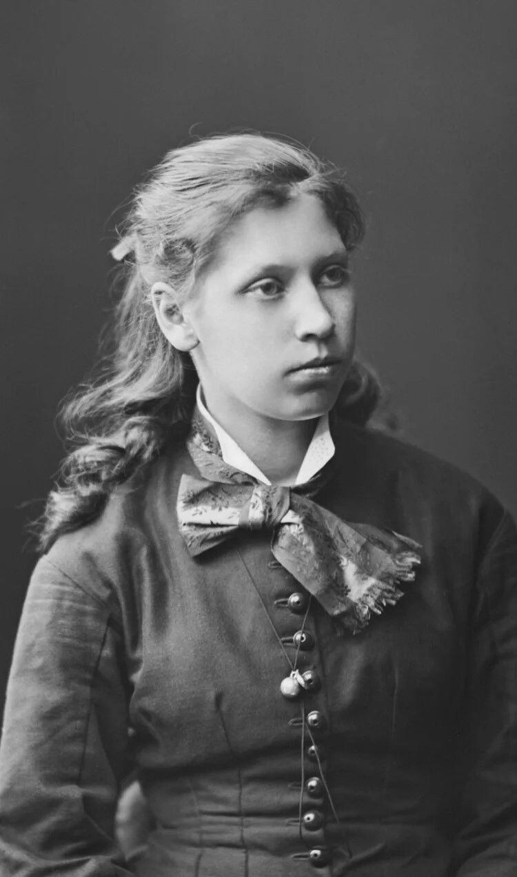 Nainen, jolla on pitkät laineikkaat hiukset, tumma napitettu mekko ja suuri kuvioitu rusetti.