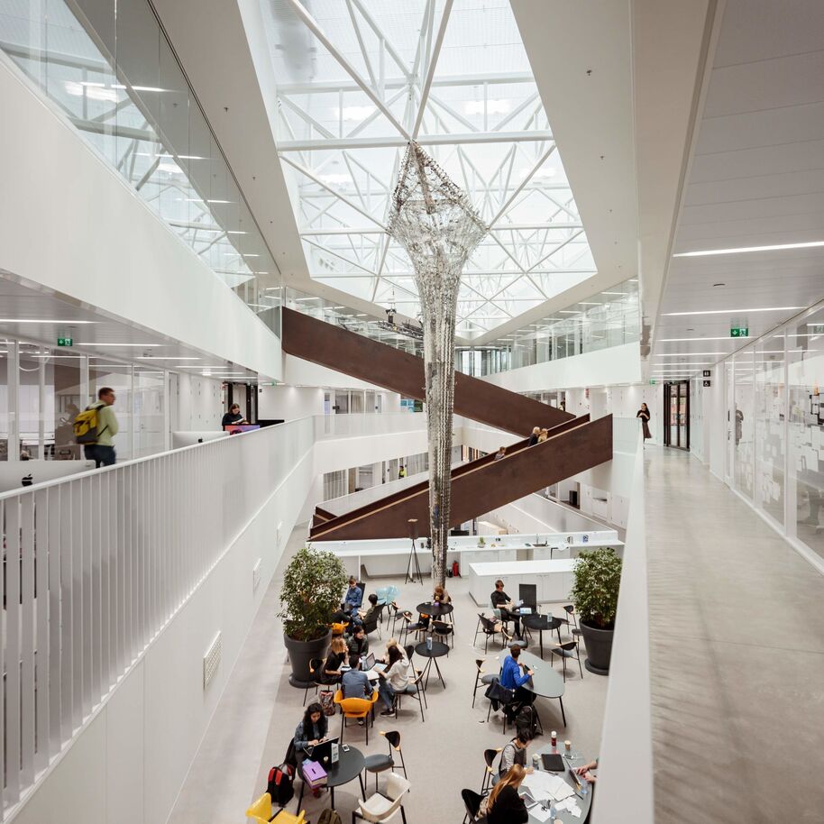 Moderni atrium, jossa suuri kattoikkuna, ruskeat siksak-portaat ja ihmisiä työskentelemässä pöydissä. Vihreät kasvit tuovat luontoa.