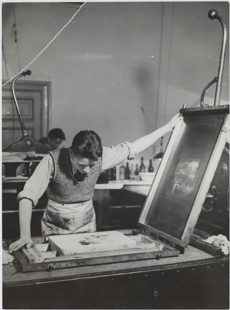 Henkilö työskentelee ison painokoneen kanssa työpajassa, yllään liivi ja esiliina.