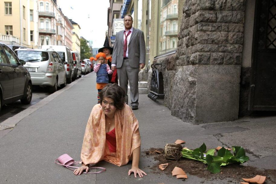 A woman in a pink dress and shawl is on the pavement next to a broken plant pot. A man and child are in the background.