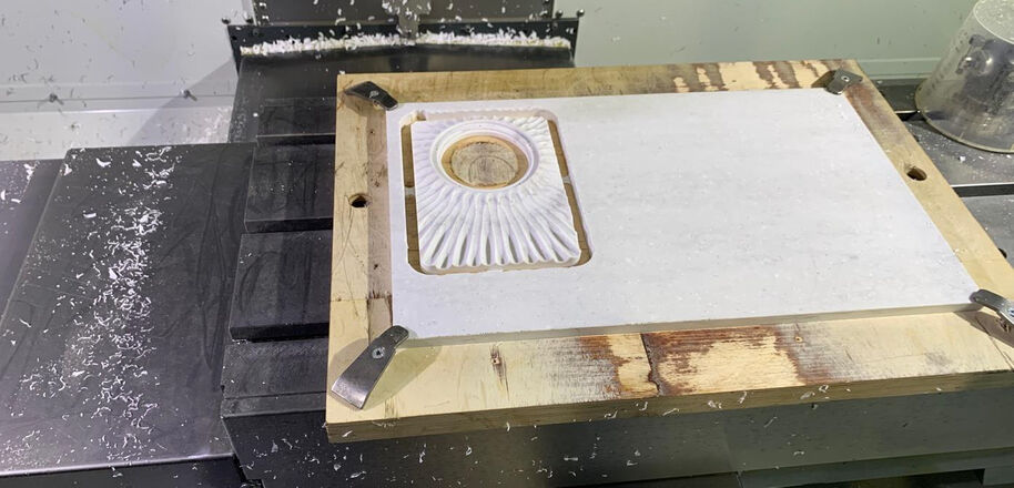 A CNC machine carving a white material with a circular pattern. Wood frame and metal clamps hold the material in place.