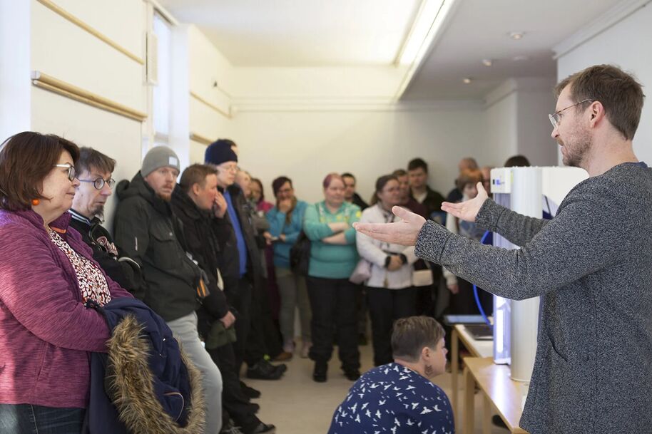 A man in a grey sweater speaks to a group of people in a room. Some people are standing, others are seated.