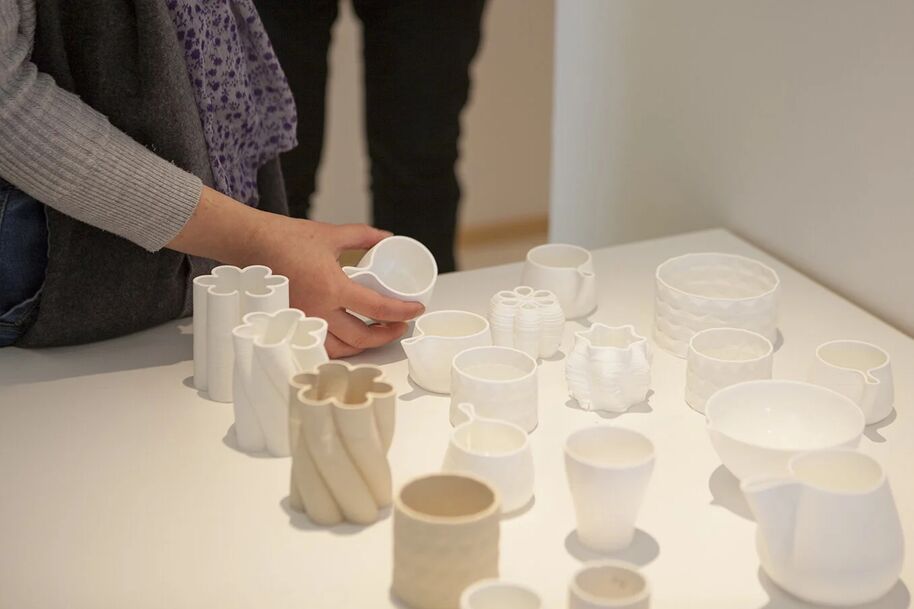 A person arranges white ceramic cups and bowls on a table. Another person stands in the background.