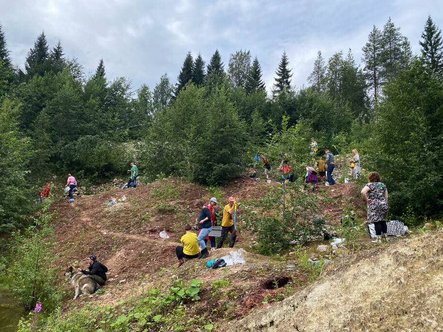 People are digging on a hillside surrounded by trees. Some are carrying buckets and tools. A dog is sitting nearby.