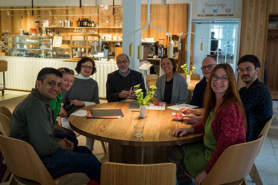A group of people sitting around a wooden table in a cafe, with notebooks and plants on the table.