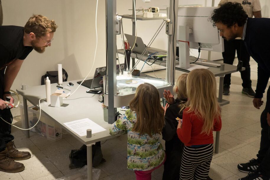 Adults and children observe a table with scientific equipment, laptops, and papers in a room with tiled flooring.