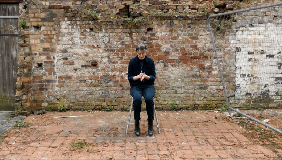 A person sits on a chair in front of a weathered brick wall in an outdoor area with a metal fence to the right.