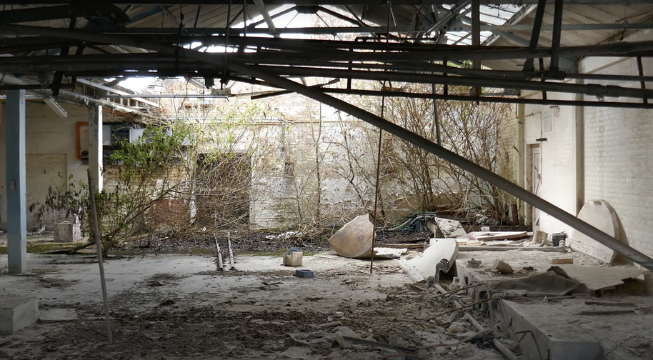 An abandoned building with debris and overgrown plants. Broken roof beams and scattered objects are visible.