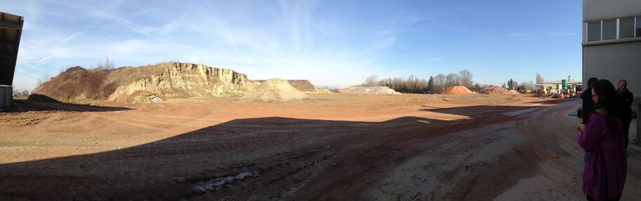 A barren construction site with dirt piles and a rocky hill. People stand on the right side near a building.