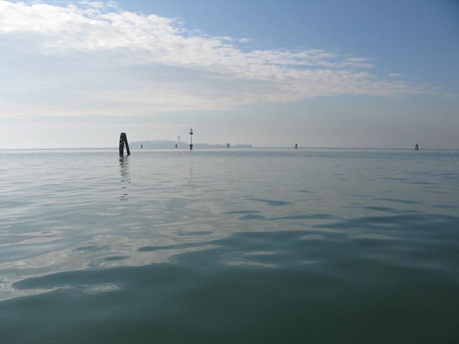 Calm sea with wooden posts and a distant shore under a partly cloudy sky.