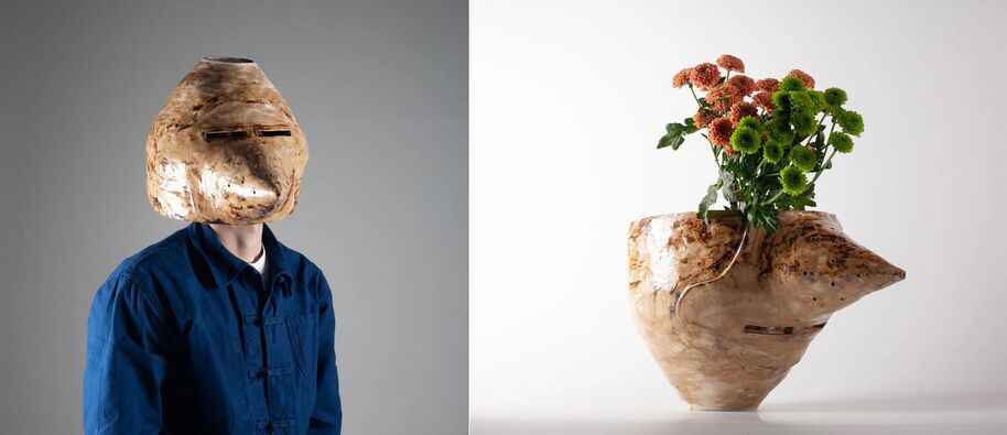 A person wearing a large, beige mask and a blue shirt next to a beige vase with green and orange flowers.