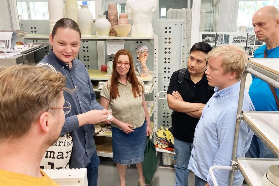 A group of people stand in a workshop with shelves holding pottery and sculptures. Some are engaged in conversation.