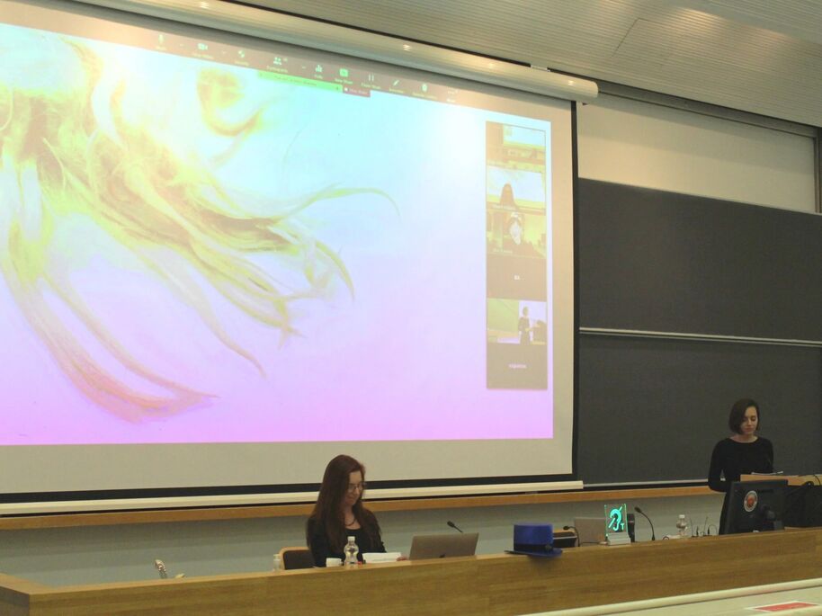 Two people in a lecture hall, one seated and one standing, with a large screen displaying a colourful image and video call participants.