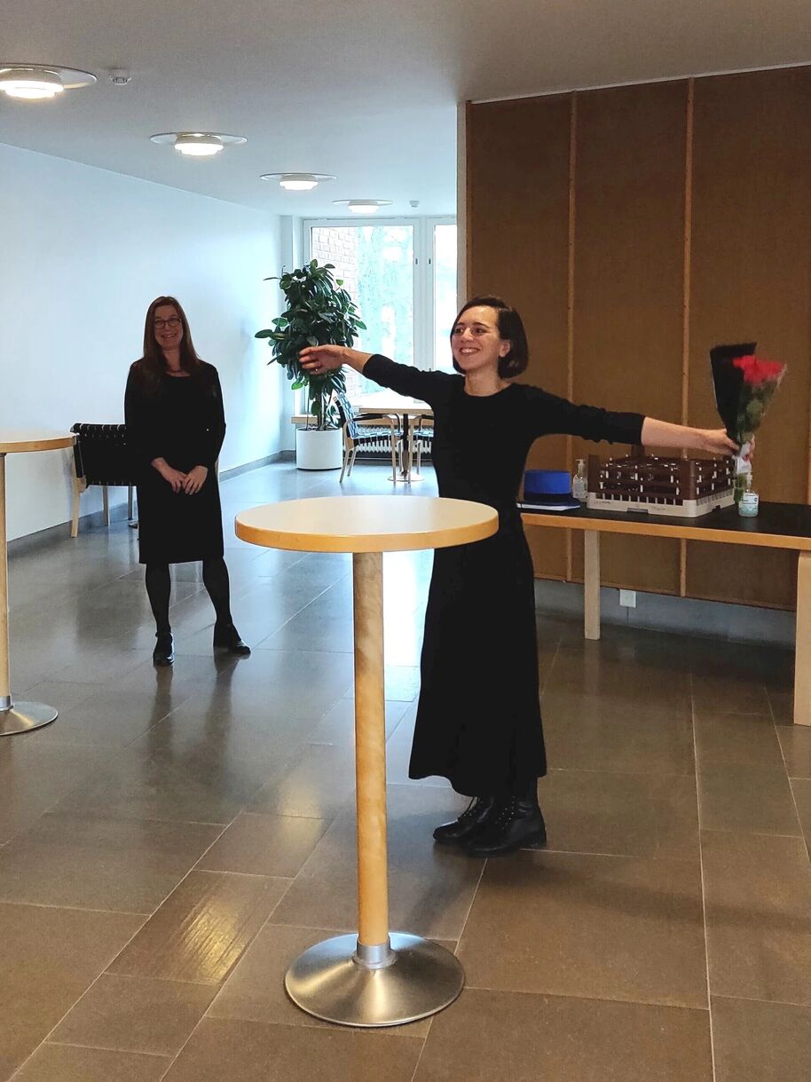 Two people in black outfits, one holding flowers, in a room with tables and a large plant near the window.