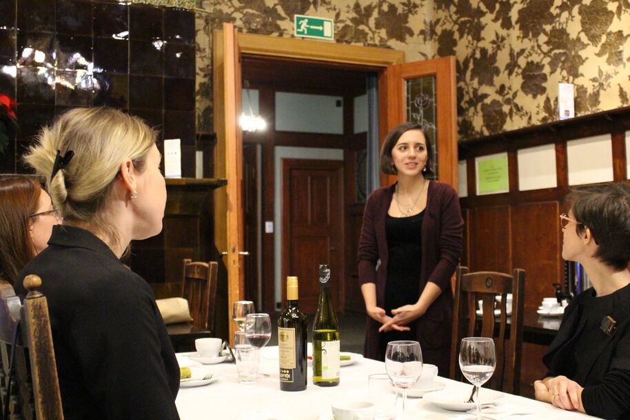 A group of people are seated at a table with wine bottles and glasses, listening to a standing woman in a brown room.