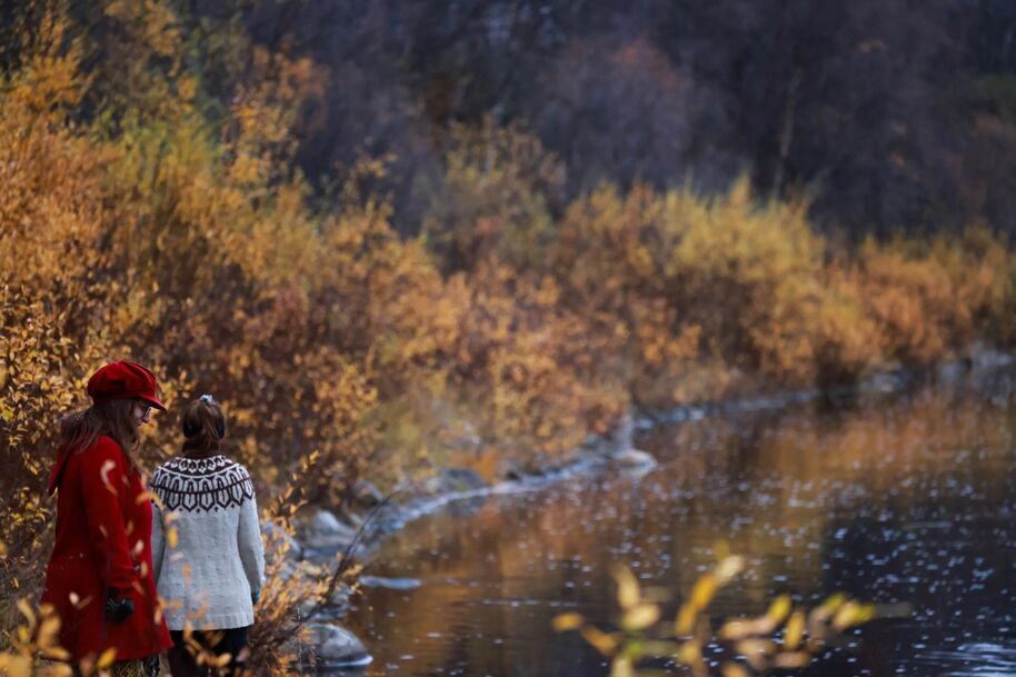 Two people walking by a river in autumn. One wears a red coat and hat, the other a white sweater with patterns.