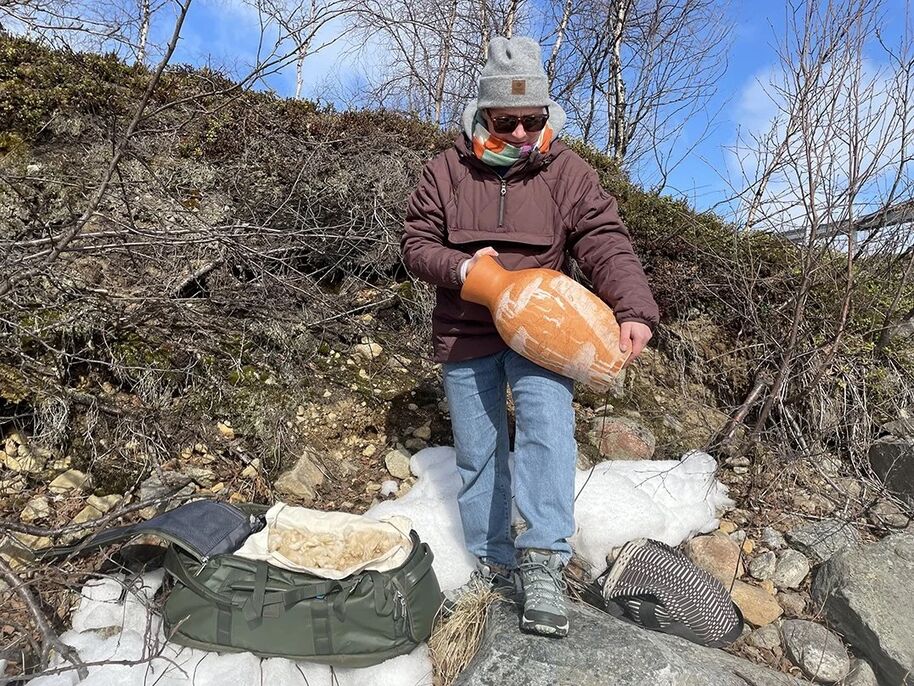 Person holding a large ceramic jug outdoors, surrounded by rocks and snow, with a green bag and striped cloth nearby.
