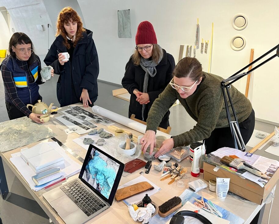 Four people are gathered around a table with art supplies, a laptop, and various objects. One person is pouring tea.