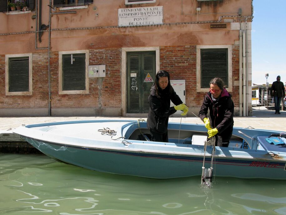 Two people on a boat collecting water samples near a brick building with green shutters.