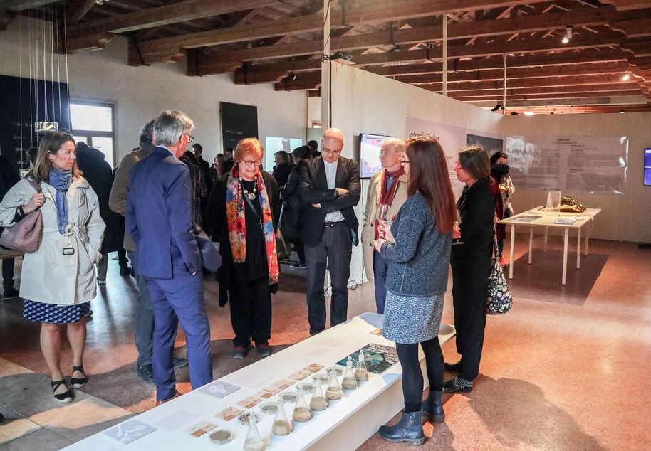 People in a gallery space with wooden beams, discussing exhibits on tables. Some hold drinks, and one wears a colourful scarf.